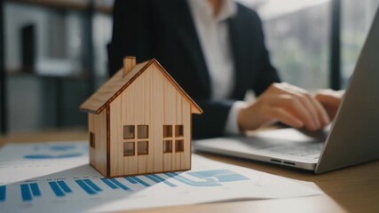 A person working on a laptop with a wooden house model and financial documents on a desk, symbolizing real estate business. - Powered by Adobe