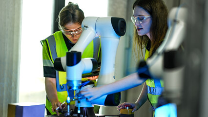 Two female engineers in safety vests and goggles collaborate in a tech lab. They are inspecting and programming a high tech robotic arm for a new project.