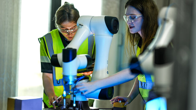 Two female engineers in safety vests and goggles collaborate in a tech lab. They are inspecting and programming a high tech robotic arm for a new project.