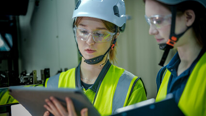 Two female engineers in hard hats and safety vests collaborate in a factory. One woman smiles and touches a control panel while her colleague holds a tablet.