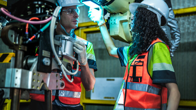Diverse engineering team working with a white industrial robotic arm. A woman with curly hair in safety gear is inspecting the machine part, while a male colleague holds a digital control panel. The s - Powered by Adobe