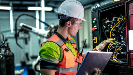 Side view of a young male engineer in a white helmet and orange safety vest checking electrical wires in a black control panel. He holds a digital tablet in one hand and touches the equipment with his