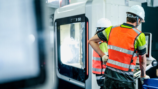 A diverse team of engineers in hard hats and safety vests operates a high-tech machine. They are programming the advanced equipment in a futuristic smart factory. - Powered by Adobe