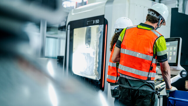 A diverse team of engineers in hard hats and safety vests operates a high-tech machine. They are programming the advanced equipment in a futuristic smart factory. - Powered by Adobe