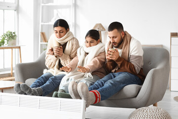 Frozen family with tea and lack of heating warming near electric convector heater at home