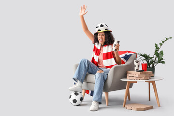 Female African-American soccer fan with drink and UK flag in armchair watching game on light background