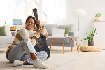 Happy young African-American woman with acoustic guitar sitting on floor in living room