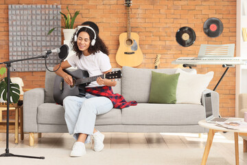 Obraz premium Pretty young African-American woman in headphones with microphone playing acoustic guitar and singing on sofa in living room