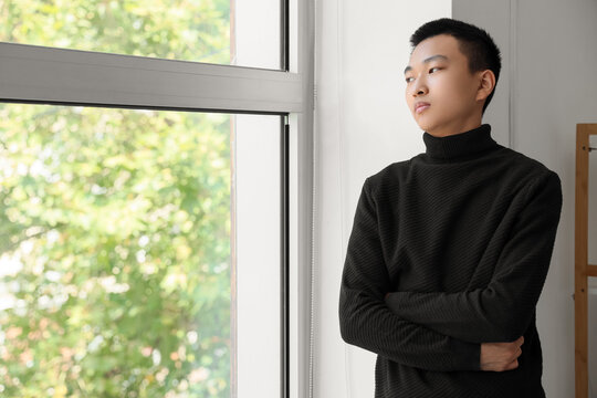 Sad young Asian man near window at home