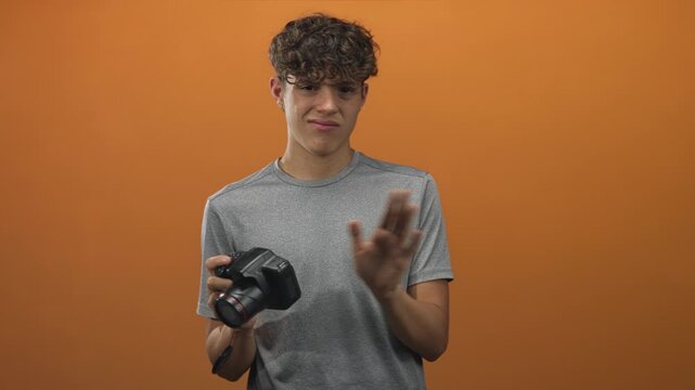 Young man holding a dslr camera with lens cap dangling and hand raised in a studio wearing gray shirt and making a wary grimace; hesitation doubt.