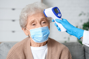 Female doctor measuring temperature of elderly woman in medical mask in living room