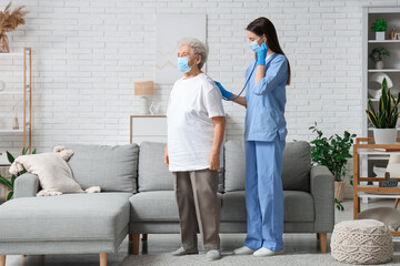 Female doctor with stethoscope examining elderly woman in medical mask in living room