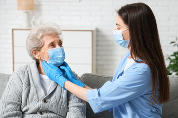 Female doctor examining elderly woman in medical mask on sofa in living room