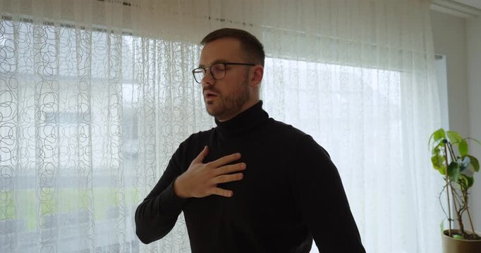 Calm lifestyle shot of a man practicing deep breathing with a hand on his chest near a bright window&mdash;concept for anxiety relief, mindfulness, self-care, and emotional regulation.