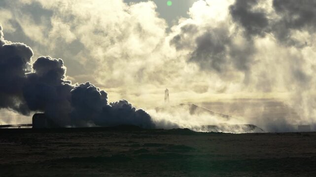 Stable shot of geothermal smoke moving across the shot into the wind while lighthouse on top of a hill becomes visible in the background.