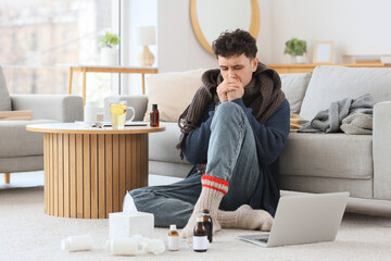 Ill young man with bottles of cough syrup and modern laptop sitting on carpet in living room