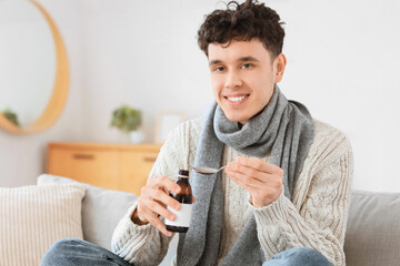 Ill young man with spoon and bottle of cough syrup sitting on sofa in living room