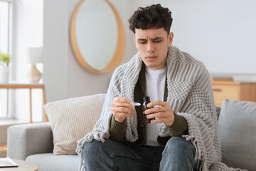 Ill young man with bottle of cough syrup and measuring spoon sitting on sofa in living room