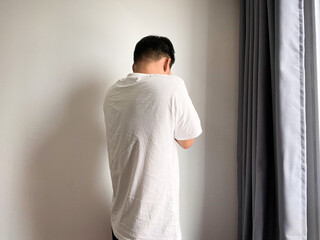 A young man in a white t-shirt standing against a white wall, bowing his head and covering his face with his hands in a gesture of sadness