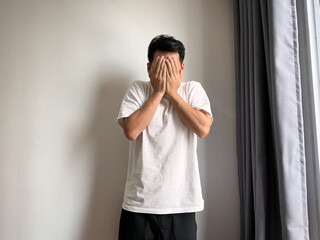 a young Asian man standing against a plain white wall, covering his face with both hands in a gesture of shame, sadness, or despair