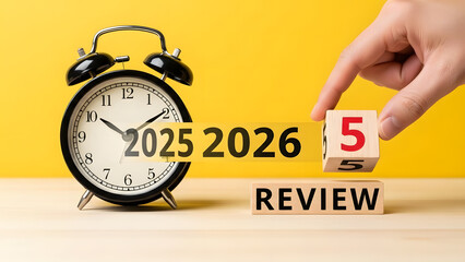A human hand places a numerical wooden block next to a classic alarm clock and "2025 2026 REVIEW" text, signifying an annual assessment or planning process.