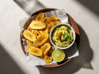 A plate of crispy fried plantain chips served with a bowl of creamy guacamole dip and a slice of lime on a white marble surface
