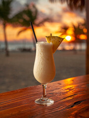 Tropical Pina Colada cocktail with pineapple slice on wooden table at sunset beach background. Refreshing summer drink in golden hour light with palm trees silhouettes and ocean view.