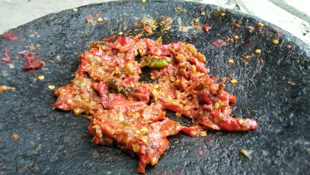 Close-up of homemade Sambal Ulek (Indonesian chili paste) freshly ground on a traditional dark stone mortar (cobek), showing spicy red chili peppers and seeds.