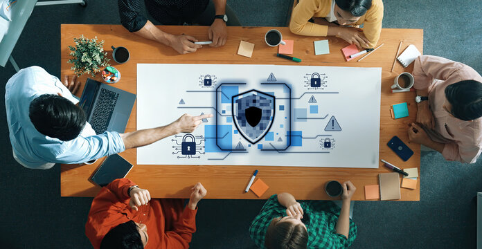 A diverse business team collaborates on a cybersecurity strategy session, utilizing a digital interface featuring a security shield and various risk alerts. Trope - Powered by Adobe
