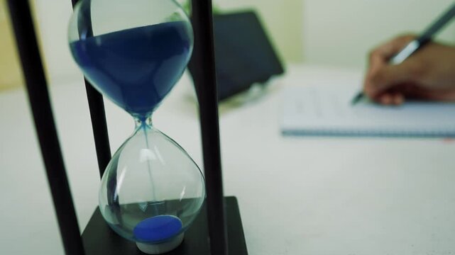 Hourglass with blue sand and person writing in notebook