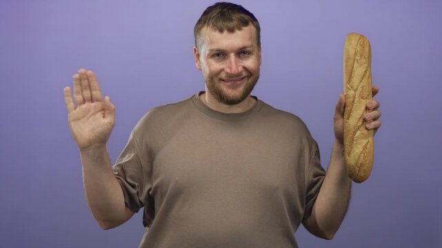 Man waves hand and holds baguette in purple studio set wearing casual brown t shirt, smiling at camera; cheerful greeting.