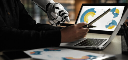 A robotic hand engages with a human partner, highlighting a collaborative effort on data analysis displayed on a laptop, showcasing modern workplace synergy.Noogenesis
