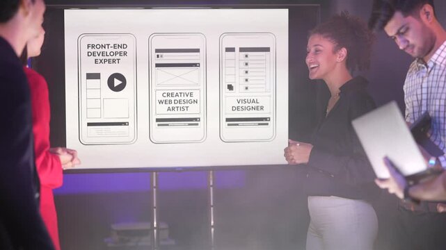 A smiling woman with curly hair discussing mobile app wireframes on a large presentation screen with a male colleague holding a laptop. The screen displays sketches for "Front-End Developer Expert" an