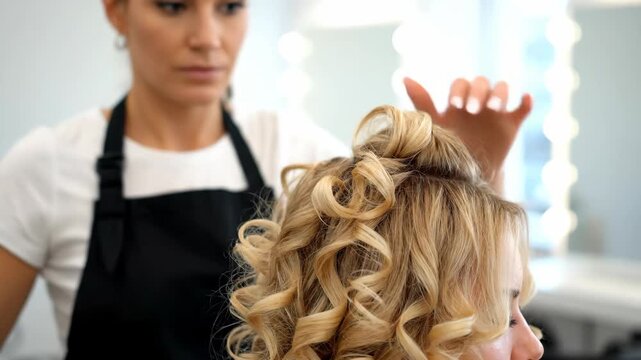 A woman's hair is styled with curls by a professional hairstylist in salon. The hairstylist touches the blonde hair