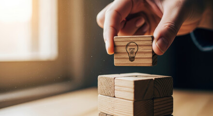 A person's hand places a wooden block with a lightbulb symbol on top of stacked blocks on a table indoors.