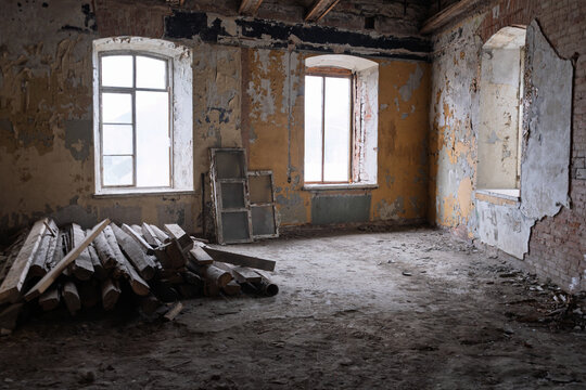 Derelict prison interior featuring two arched windows, crumbling walls with peeling yellow paint. Abandoned jail room with debris pile, exposed wooden beams, damaged plaster, and atmospheric decay