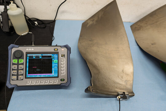 Ultrasonic NDT inspection equipment for testing aircraft engine fan blades with a handheld flaw detector displaying scan data on a digital screen during aerospace quality control and defect detection