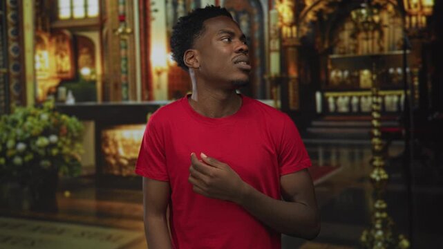 Young african american man in red t shirt raises hand while looking upward inside church building; devotion.