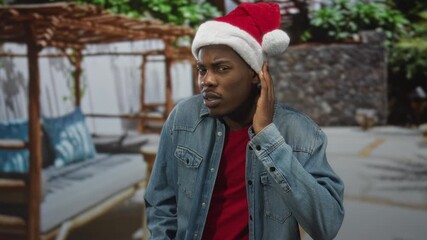 Man wearing santa hat and denim jacket by building, hand to ear listening gesture; holiday curiosity wonder.
