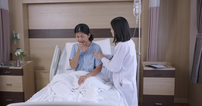 Asian female patient sitting on hospital bed coughs during consultation as female doctor checks condition and provides medical advice for respiratory issue and ongoing treatment