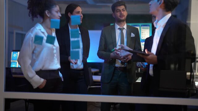 A young businessman in a suit writing on a glass wall with a green marker during a brainstorming session. A woman with curly hair stands nearby looking at blue sticky notes. Colleagues in the backgrou