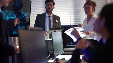 Diverse business group having a discussion in a dark blue-lit room. A man in a suit holding a tablet stands next to a woman with curly hair holding papers. They are presenting to colleagues with a dig - Powered by Adobe