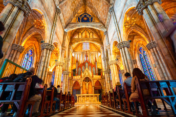 Obraz premium Grand church interior featuring intricate Gothic architecture, vibrant stained glass windows, and people reverently seated in wooden pews.