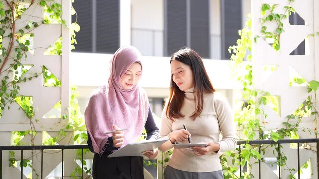 Two businesswomen, one wearing a hijab, standing in ffice building corridoor, smiling and talking while holding clipboard document and tablet.