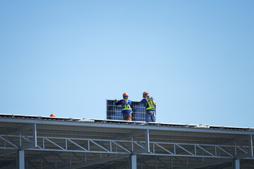 Two worker who wearing full safety PPE are installing solar cell panel on the factory rooftop with blue sky background. Industrial working scene.  © Nattawit