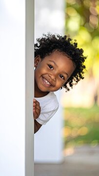 Joyful young girl with curly hair peeking around a white corner outdoors.