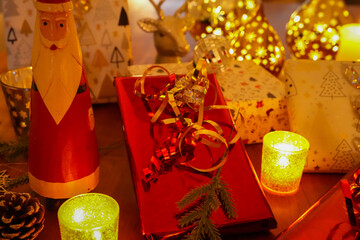 Festive Christmas decoration with several wrapped gifts, a traditional Santa Claus figure, pine cones and burning candles on a wooden surface