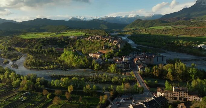 Drone glide approaches A&iacute;nsa from the Mirador de Fuebla side, flying above the R&iacute;o Cinca channels, riverside woodland toward fortified medieval village and castle on hill with snowy Pyrenees peaks.