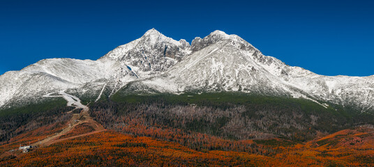 Tatranske Matliare, Slovakia - Aerial panoramic view of the snowy mountains of Lomnicky Peak in the High Tatras with red and orange coloured autumn trees and foliage and clear blue sky at Vysoke Tatry