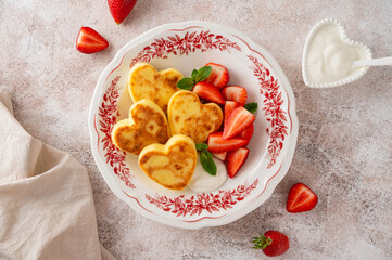 Heart-shaped cottage cheese pancakes or syrnyky with sour cream and fresh strawberries on a plate on a concrete background. Breakfast for Valentine's Day or Mother's Day. Top view, copy space.